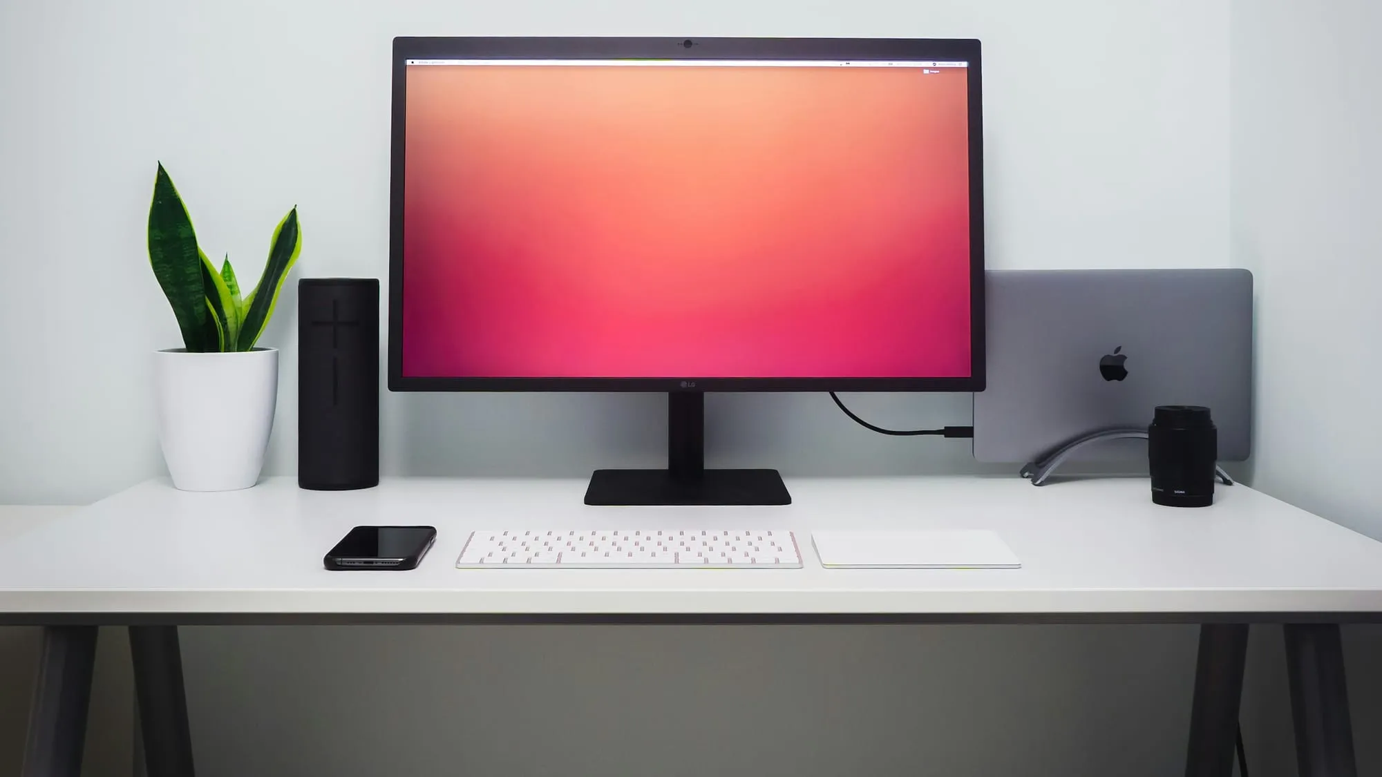 A monitor, keyboard and trackpad on a white desk against a pale wall, with a plant and a speaker beside the screen